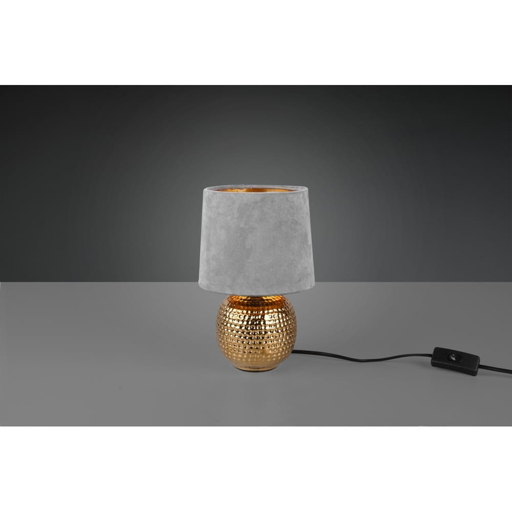 Szara lampa stołowa Sophia z 1 punktem świetlnym, posiadająca okrągły, złoty podstawę z teksturowanej powierzchni oraz miękki, szary klosz z materiału. Produkt stoi na stole, z widocznym przewodem z włącznikiem.