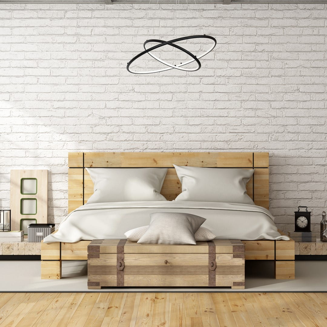 Czarna lampa wisząca Orion LED 2-punktowa w nowoczesnym stylu, zawieszona nad łóżkiem, z dwoma okrągłymi pierścieniami o minimalistycznym designie. Tło stanowi biała ceglana ściana oraz drewniane meble w jasnym wykończeniu.