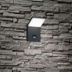 Kinkiet elewacyjny Pearl LED 1-punktowy w antracytowym kolorze, z czujnikiem ruchu. Lampa montowana na ścianie, z prostokątną, lekko pochyloną osłoną z białego materiału, umieszczoną nad okrągłym czujnikiem. Tło to teksturowana ściana z kamienia.