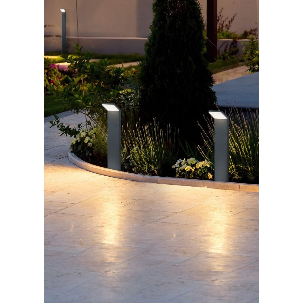 Lampa stojąca ogrodowa Pearl H50 LED w antracytowym kolorze, z prostokątnym korpusem, umieszczona na ścieżce otoczonej kwiatami. Kąt ujęcia z boku, pokazujący delikatne oświetlenie otoczenia. W tle widoczna zieleń i kamienna nawierzchnia.