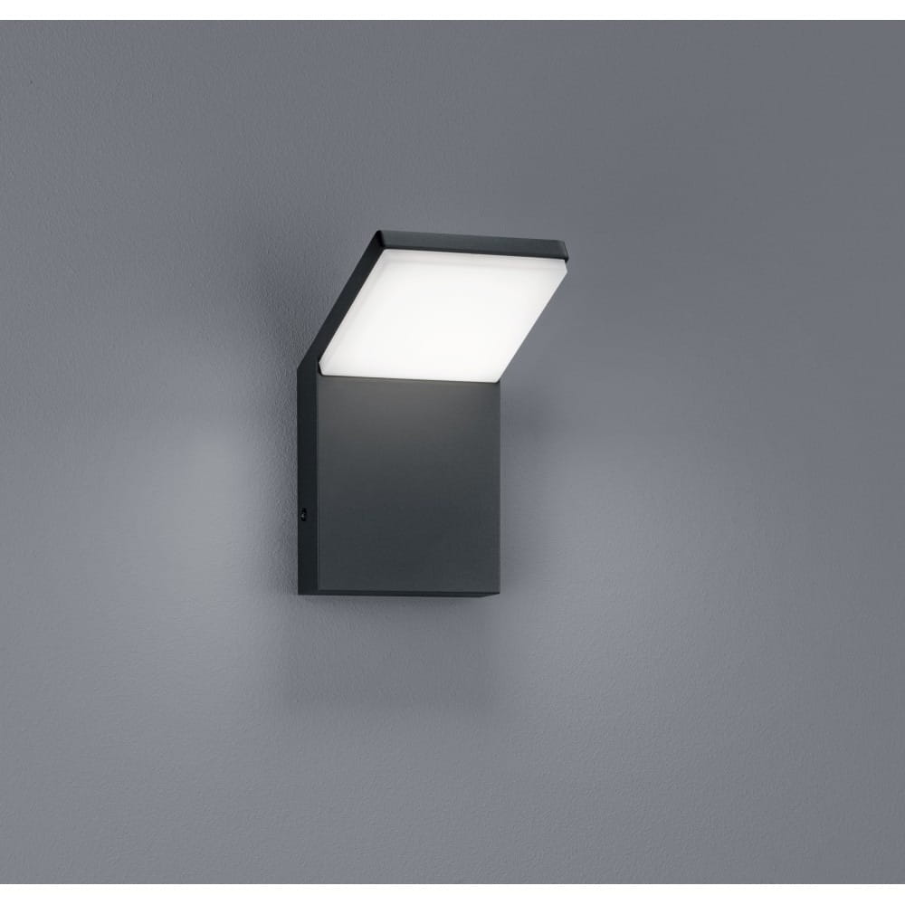 Kinkiet elewacyjny Pearl LED w kolorze antracytowym, 1-punktowy. Przesunięty klosz w kształcie prostokąta, zamontowany na ścianie. Emituje światło w dół, z prostą, minimalistyczną formą. Tło w odcieniu szarości.