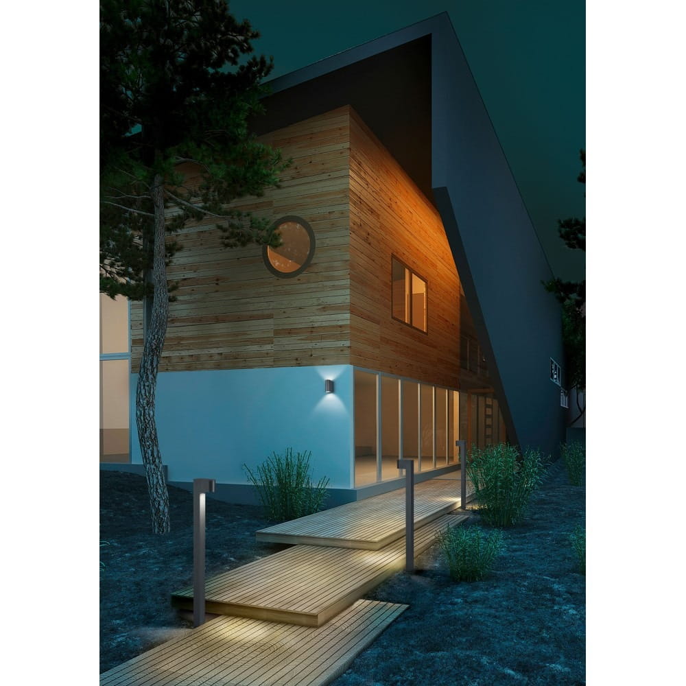 Lampa stojąca ogrodowa Roya H100, antracytowa, umieszczona obok drewnianego chodnika na tle nowoczesnego domu. Produkt ma prosty, cylindryczny kształt, emitując światło, co tworzy przyjemny nastrój w otoczeniu.