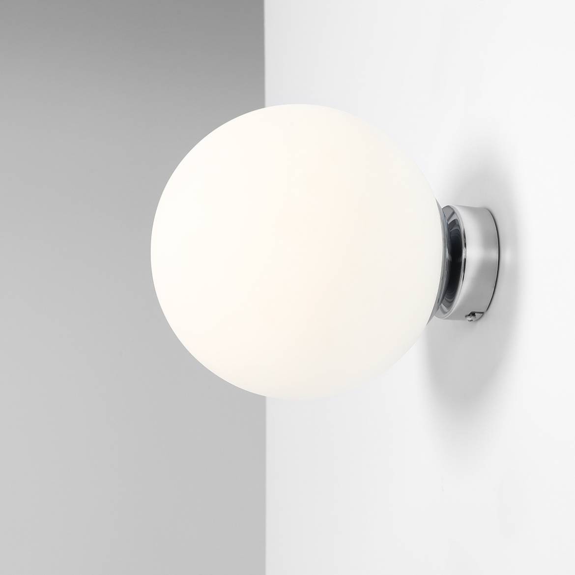 Kinkiet Ball D20 w kolorze chromu z białą, okrągłą, matową kloszem. Zamontowany na ścianie, widoczny z boku. Prosta forma, idealny do nowoczesnych wnętrz. Tło w odcieniach szarości, co podkreśla elegancję lampy.
