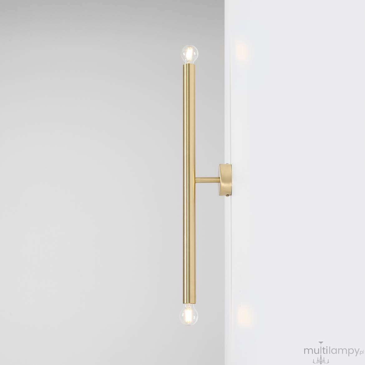 Kinkiet Tubo 2-punktowy w kolorze mosiądzu, z długim, prostym ramieniem i dwoma oprawkami na żarówki. Zainstalowany na białej ścianie, prezentuje minimalistyczny design. Widoczny kształt cylindryczny, metaliczna faktura.