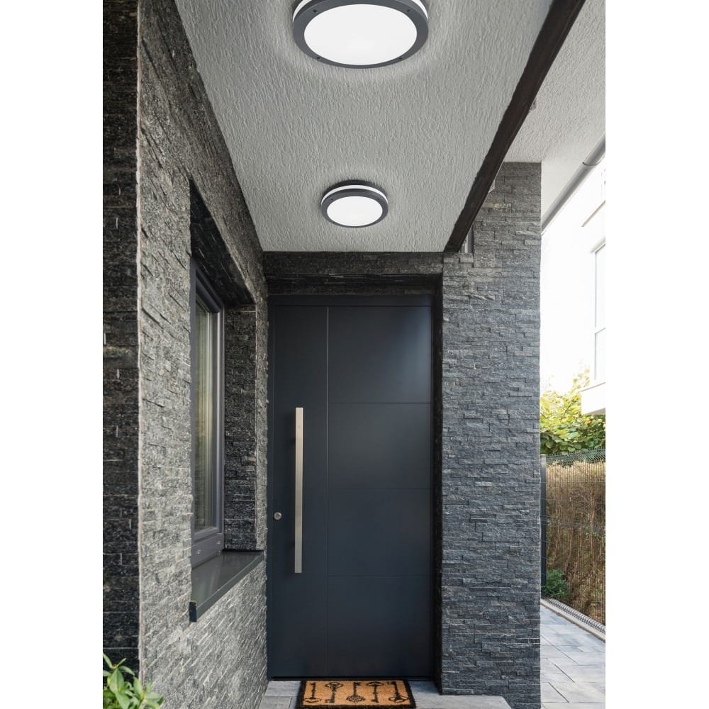 Piave LED lampa sufitowa zewnętrzna, antracytowa, z czujnikiem ruchu. Ujęcie z boku, montowana pod sufitem. Widoczne dwa okrągłe elementy o matowej, białej powierzchni. Lampa zamontowana w eleganckim otoczeniu przed wejściem do budynku.