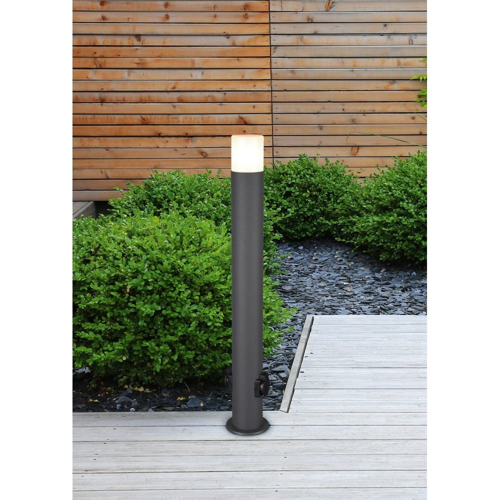 Lampa stojąca Hoosic H80 w kolorze antracytowym, z okrągłym korpusem i mlecznym kloszem. Posiada dwa gniazda elektryczne. Ustawiona na drewnianym podłożu, otoczona zielonymi krzewami, w tle drewniana ściana.