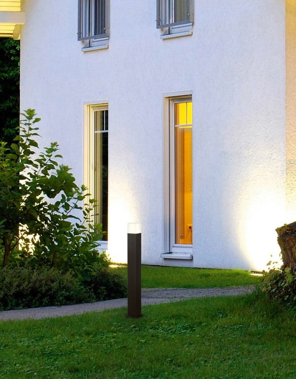 Lampa stojąca ogrodowa Hudson H80 w kolorze antracytowym, o cylindrycznym kształcie, umieszczona na trawniku przed białą ścianą budynku. Górna część lampy emituje światło, oświetlając otoczenie. W tle widoczne okna oraz roślinność.