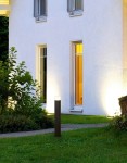 Lampa stojąca ogrodowa Hudson H80 w kolorze antracytowym, o cylindrycznym kształcie, umieszczona na trawniku przed białą ścianą budynku. Górna część lampy emituje światło, oświetlając otoczenie. W tle widoczne okna oraz roślinność.