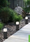 Lampa stojąca ogrodowa Hudson H30 w kolorze antracytowym, z prostokątną podstawą i kwadratowym kloszem. Ujęcie z boku, pokazujące lampy wzdłuż ścieżki, otoczone roślinnością i żwirem. Idealna do oświetlenia przestrzeni zewnętrznych.