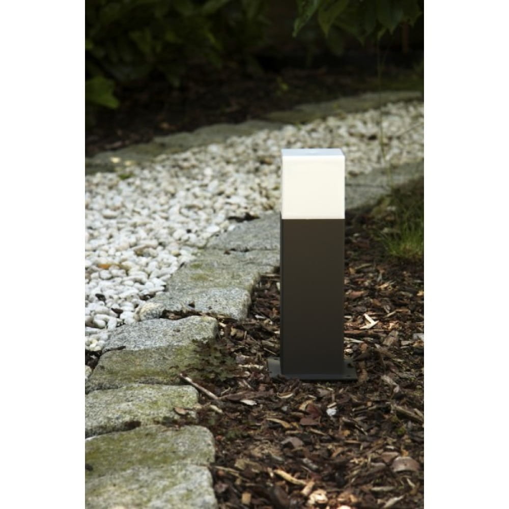 Lampa stojąca ogrodowa Hudson H30 w kolorze antracytowym, jednostkowa, o prostokątnym kształcie. Górna część biała, dolna antracytowa. Ustawiona na tle żwiru i kory, w otoczeniu roślin.