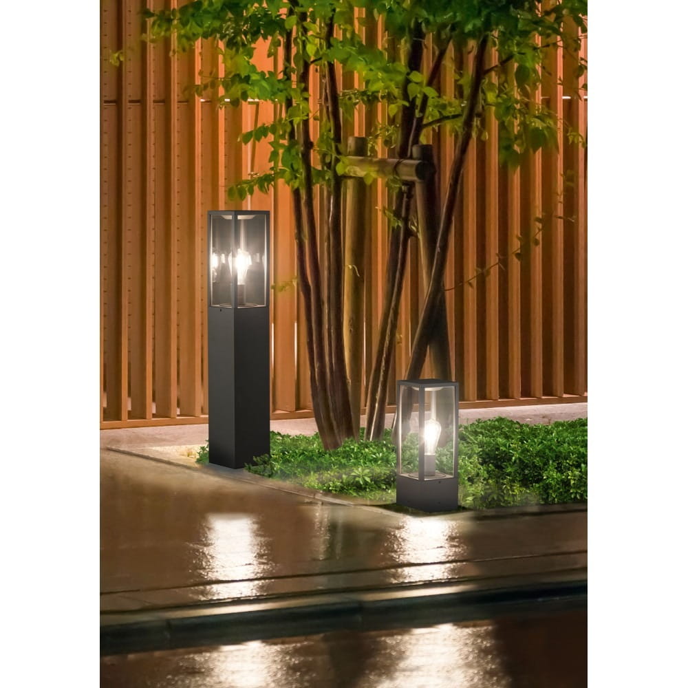 Lampa ogrodowa stojąca Garonne H40 w kolorze antracytowym, z prostokątną podstawą, oświetleniem w górnej części. Umieszczona na mokrej nawierzchni, w otoczeniu roślinności, z drewnianym tłem. Widać również mniejszą lampę obok.