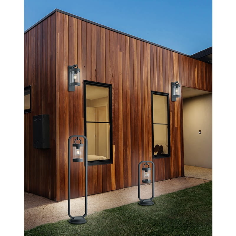 Kinkiet zewnętrzny Sambesi 1-punktowy w kolorze antracytowym z czujnikiem zmierzchu, zamontowany na drewnianej ścianie budynku, obok dwóch lamp stojących. Widoczna metalowa konstrukcja oraz szklane klosze. Otoczenie trawnika i podjazdu.