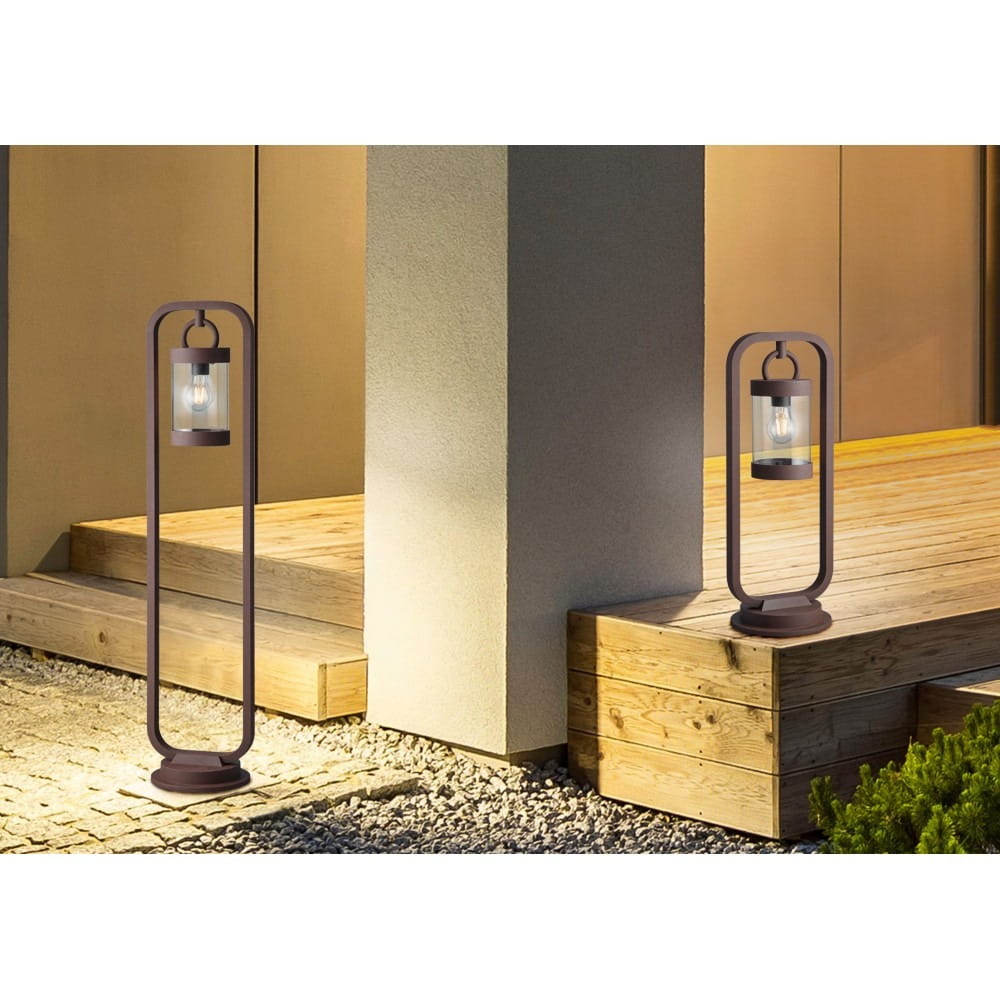 Lampa ogrodowa Sambesi H100 w kolorze rdzawej, stojąca, 1-punktowa z czujnikiem zmierzchu. Zawieszona w metalowej ramie, umieszczona na tle drewnianego tarasu i jasnej ściany, emitująca ciepłe światło.