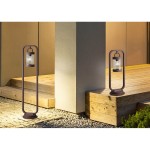 Lampa ogrodowa Sambesi H100 w kolorze rdzawej, stojąca, 1-punktowa z czujnikiem zmierzchu. Zawieszona w metalowej ramie, umieszczona na tle drewnianego tarasu i jasnej ściany, emitująca ciepłe światło.