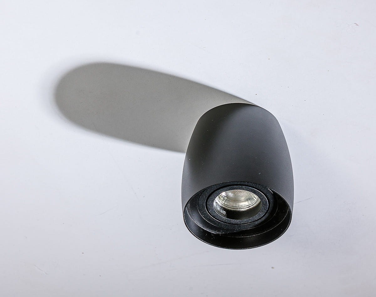 Rotondo lampa sufitowa kierunkowa w kolorze czarnym, o zaokrąglonym kształcie. Ujęcie z góry, pokazujące reflektor z widocznym wnętrzem. Lampa montowana do sufitu, z delikatnym cieniem na tle.