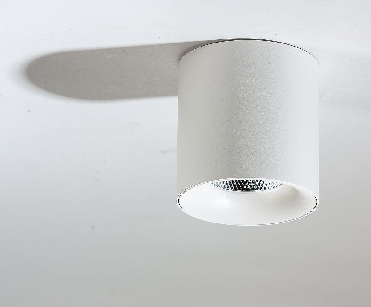 Biała lampa sufitowa LED 1-punktowa o mocy 10W i temperaturze barwowej 4000K. Cylindryczny kształt, matowa powierzchnia, z widoczną soczewką w centralnej części. Zamontowana na suficie, rzucająca delikatny cień.