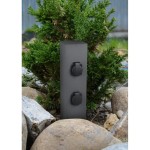 Słupek ogrodowy antracytowy z dwoma wbudowanymi gniazdami z uziemieniem, stojący wśród kamieni i roślinności. Widoczny prostokątny kształt z zamkniętymi pokrywami gniazd.