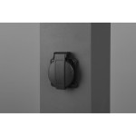 Słupek ogrodowy w kolorze antracytowym z wbudowanym gniazdem z uziemieniem. Ujęcie z boku, pokazujące gniazdo zamontowane na powierzchni słupka. Gniazdo ma czarną osłonę i oznaczenie IP54. Tło jednolite, szare.