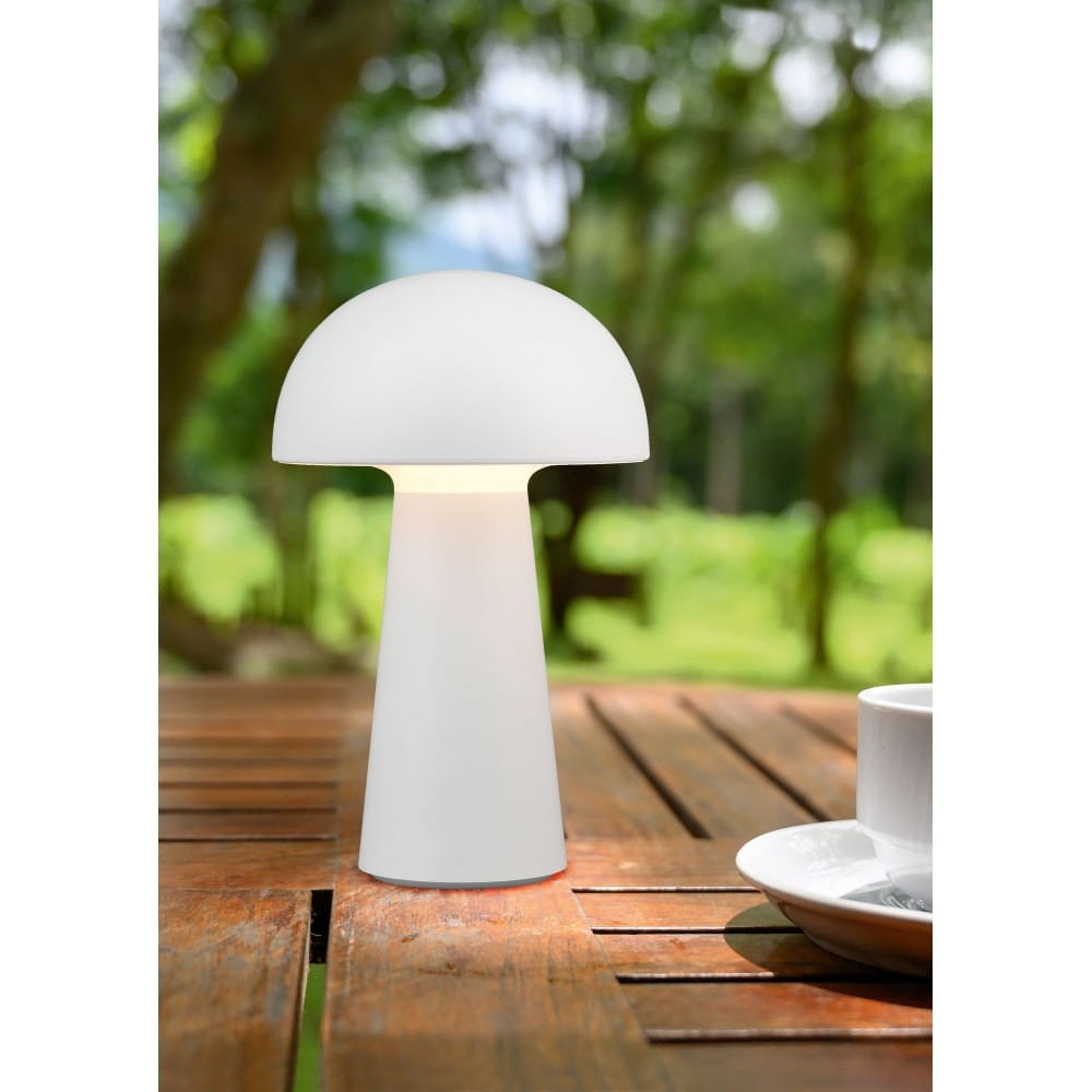 Biała lampa stołowa LED Lennon z kształtem grzyba, stojąca na drewnianym stole. Ma okrągłą, matową głowicę i cylindryczną podstawę. Tło przedstawia zieleń ogrodu, co podkreśla jej przeznaczenie do użytku na zewnątrz.
