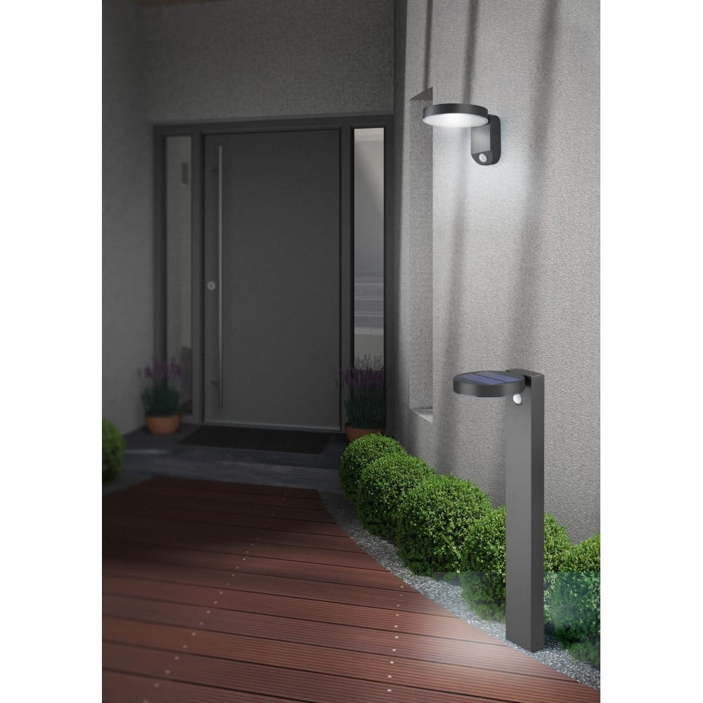 Lampa ogrodowa Posadas H60 w kolorze antracytowym, z wbudowanym czujnikiem ruchu. Ustawiona pionowo obok drzwi, z panelami słonecznymi u góry. Otoczona niskimi krzewami, na tle drewnianego podestu. Oświetlenie skierowane w dół.