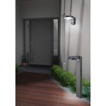 Lampa ogrodowa Posadas H60 w kolorze antracytowym, z wbudowanym czujnikiem ruchu. Ustawiona pionowo obok drzwi, z panelami słonecznymi u góry. Otoczona niskimi krzewami, na tle drewnianego podestu. Oświetlenie skierowane w dół.