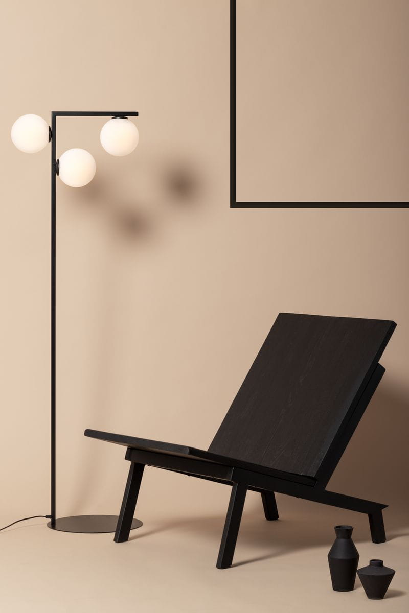 Czarna lampa podłogowa Andy z trzema okrągłymi kloszami w białym kolorze, umieszczona obok ciemnego, nowoczesnego fotela. Lampa ma prostą, metalową konstrukcję z okrągłą podstawą, na tle jasnobeżowej ściany.