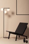 Czarna lampa podłogowa Andy z trzema okrągłymi kloszami w białym kolorze, umieszczona obok ciemnego, nowoczesnego fotela. Lampa ma prostą, metalową konstrukcję z okrągłą podstawą, na tle jasnobeżowej ściany.