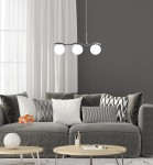 Lampa wisząca Ramon 3-punktowa w chromowanej obudowie z trzema białymi, okrągłymi kloszami. Zawieszona nad szarym sofą z poduszkami, w nowoczesnym wnętrzu z szarymi ścianami i jasnymi zasłonami.