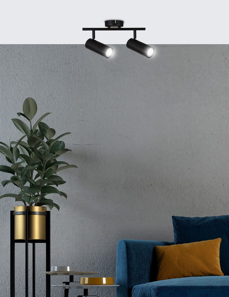 Czarna lampa sufitowa (spot) Colly z dwoma ruchomymi punktami świetlnymi, zamontowana na prostokątnej podstawie. Widok z dołu, w tle szary mur i niebieska sofa z poduszką w kolorze musztardowym, obok doniczka z rośliną.