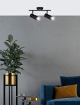 Czarna lampa sufitowa (spot) Colly z dwoma ruchomymi punktami świetlnymi, zamontowana na prostokątnej podstawie. Widok z dołu, w tle szary mur i niebieska sofa z poduszką w kolorze musztardowym, obok doniczka z rośliną.