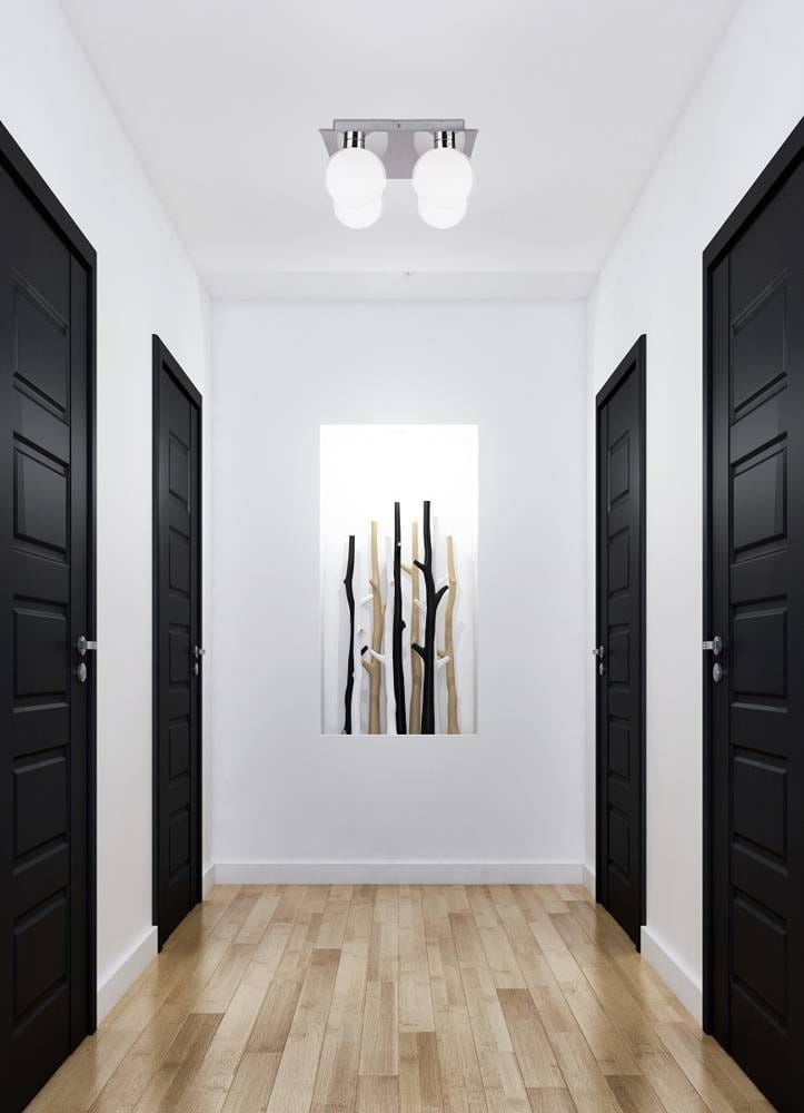Lampa sufitowa Oden 4-punktowa w chromie, z trzema białymi, okrągłymi kloszami, umieszczona na prostokątnej podstawie. Widoczna w korytarzu z ciemnymi drzwiami po obu stronach i dekoracją na ścianie.