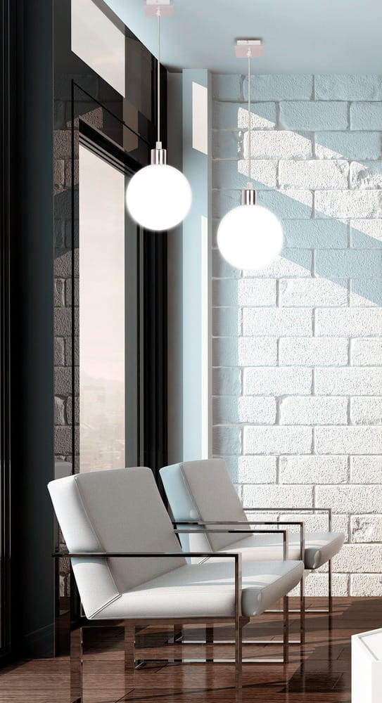 Lampa wisząca Oden D15 w chromowanej oprawie z białym, okrągłym kloszem. Zawieszona w nowoczesnym wnętrzu, obok dwóch jasnoszarych foteli na metalowych nogach. Tło z białej cegły i dużych okien, wpuszczających naturalne światło.
