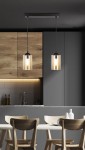 Czarna lampa wisząca Bistro z dwoma przezroczystymi kloszami, zawieszona na prostokątnej podstawie. Klosze mają złote żarówki, a lampa umieszczona jest nad drewnianym stołem w nowoczesnej kuchni z szarymi i drewnianymi elementami.