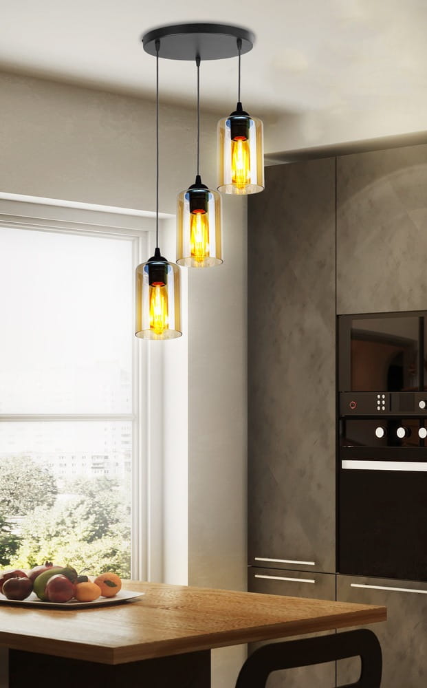 Lampa wisząca Bistro 3-punktowa w kolorze czarnym, z trzema szklanymi kloszami w kształcie cylindrów, emitującymi ciepłe światło. Zawieszona na cienkich linkach, montowana na okrągłej podstawie sufitowej, w nowoczesnej kuchni z drewnianym stołem.