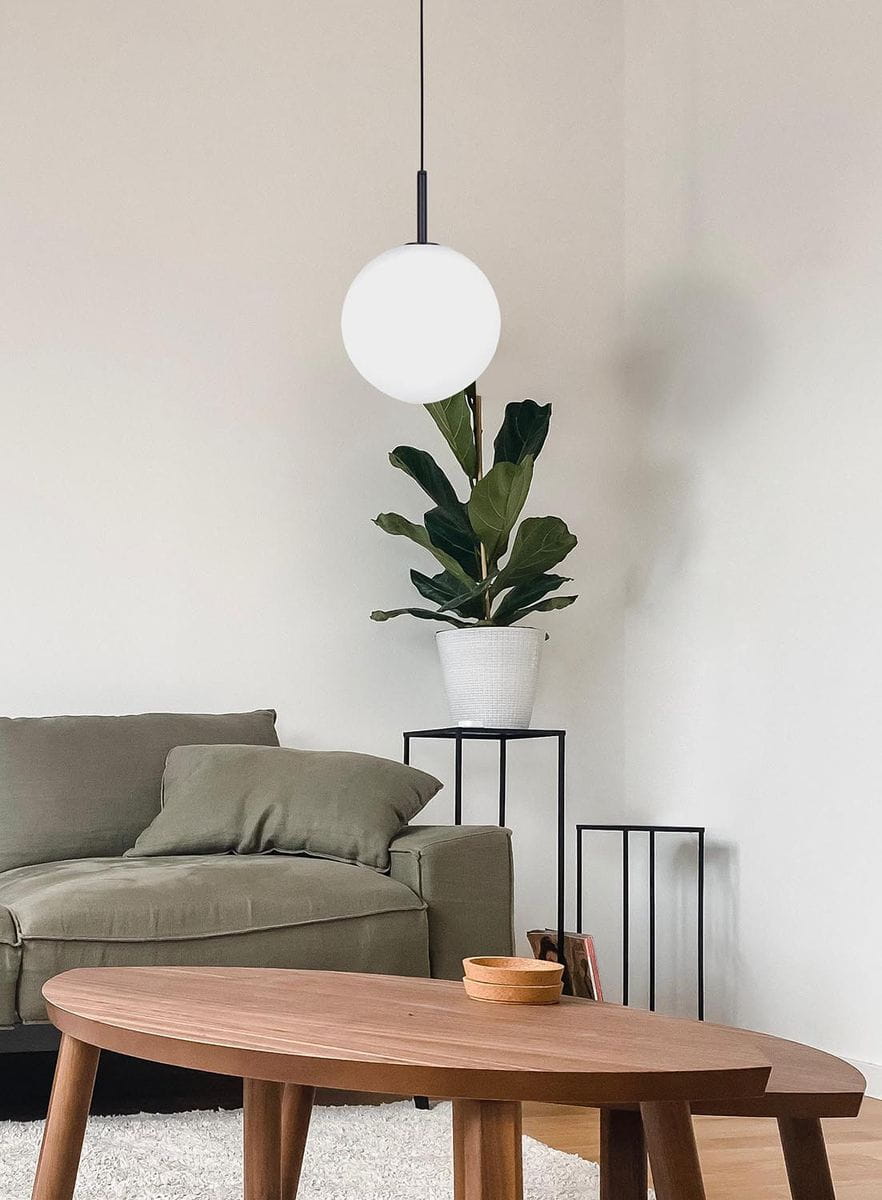 Lampa wisząca Faro D30 w kolorze czarnym, z białym, okrągłym kloszem. Zawieszona na cienkim przewodzie, w tle zielona roślina w białym doniczce oraz szara kanapa. Okrągły stół z drewnianym blatem i małym pojemnikiem. Proste, jasne tło.