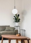 Lampa wisząca Faro D30 w kolorze czarnym, z białym, okrągłym kloszem. Zawieszona na cienkim przewodzie, w tle zielona roślina w białym doniczce oraz szara kanapa. Okrągły stół z drewnianym blatem i małym pojemnikiem. Proste, jasne tło.