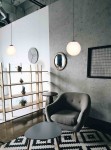 Lampa wisząca Savoy D30 w kolorze czarnym, z okrągłym, białym kloszem, zawieszona w nowoczesnym wnętrzu. Widoczna w aranżacji z szarym fotelem, czarnym stołem oraz półką z roślinami. Kąt ujęcia frontalny, z lekkim widokiem na otoczenie.