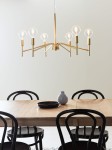 Złoty żyrandol Monarch 6-punktowy, z sześcioma przezroczystymi kloszami, zawieszony nad drewnianym stołem. Metalowa konstrukcja z minimalistycznym designem, w otoczeniu czarnych krzeseł i ceramicznych akcesoriów na stole.
