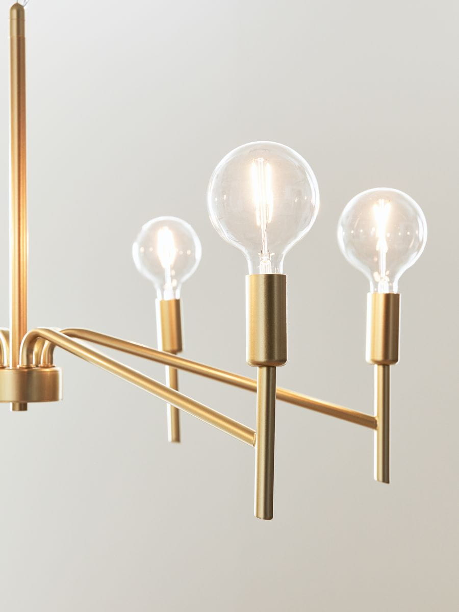 Żyrandol Monarch 6-punktowy w kolorze złotym, z przezroczystymi kloszami na żarówki. Ujęcie z boku, ukazujące trzy z sześciu punktów świetlnych. Materiał metalowy, minimalistyczny design. Tło jasne, proste.