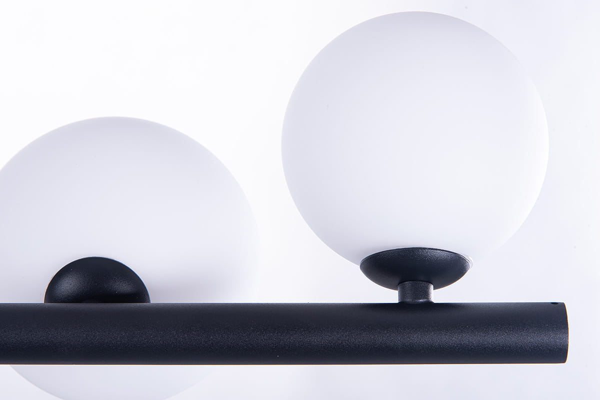 Czarna lampa wisząca Sybilla z siedmioma punktami świetlnymi. Konstrukcja z matowego metalu z białymi, okrągłymi kloszami z opalowego szkła. Ujęcie z góry, ukazujące detale mocowania kloszy i kształt lampy. Tło białe.