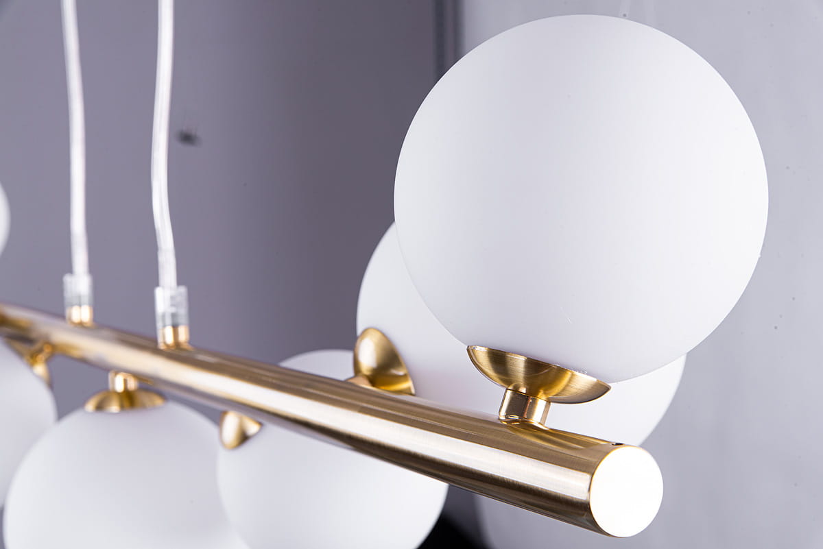 Lampa wisząca Sybilla, 10-punktowa, z metalowym, złotym stelażem i białymi, okrągłymi kloszami. Ujęcie z bliska, ukazujące detale mocowania oraz teksturę materiałów. Klosze umieszczone na poziomej listwie, zawieszone na przezroczystych przewodach.