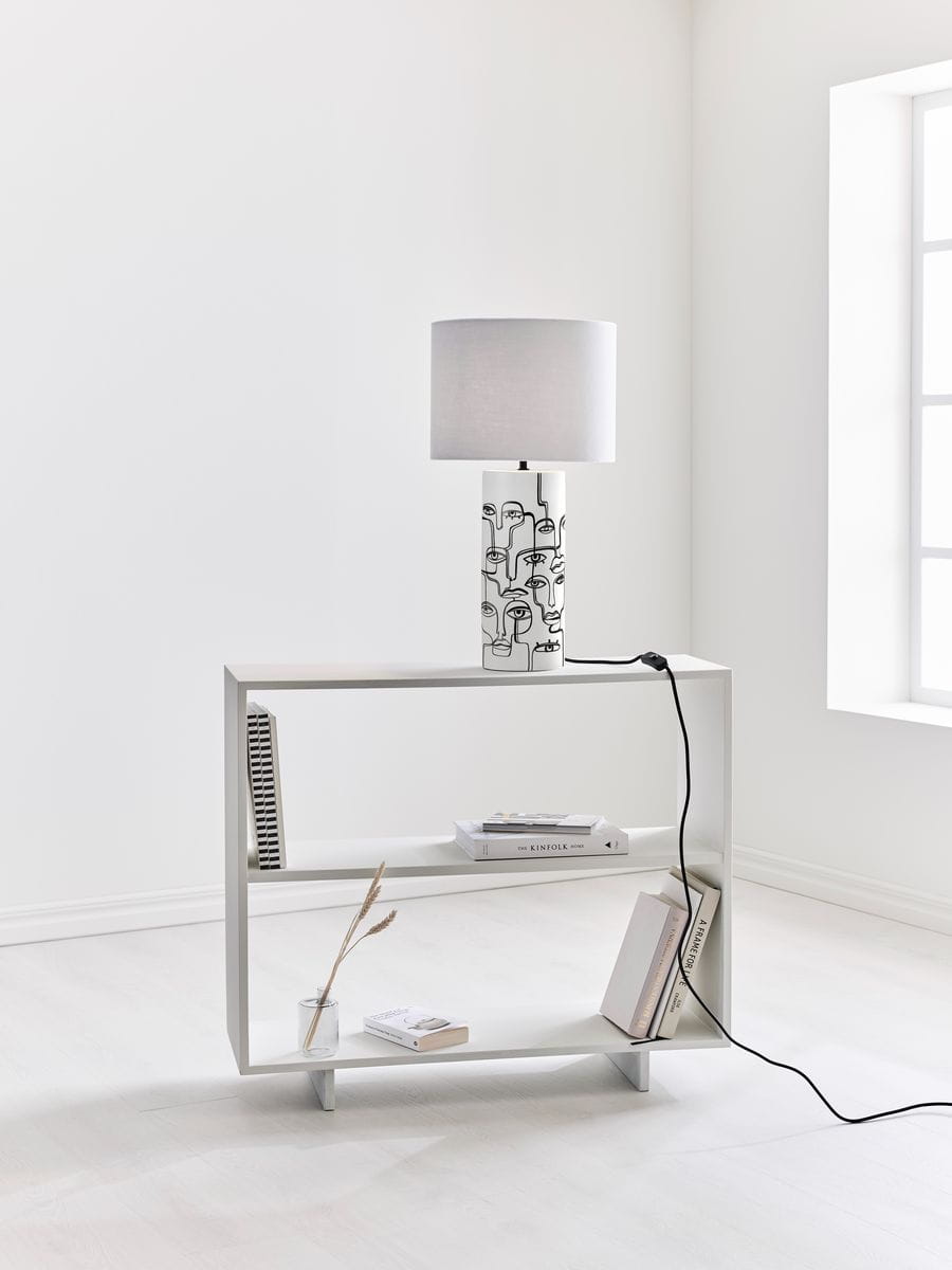 Lampa stołowa Family 1-punktowa w kolorze białym z dekoracyjnym wzorem na podstawie. Ustawiona na białej półce, z szarym kloszem. Widoczny kabel zasilający. Tło jasne, minimalistyczne, z oknem w tle.