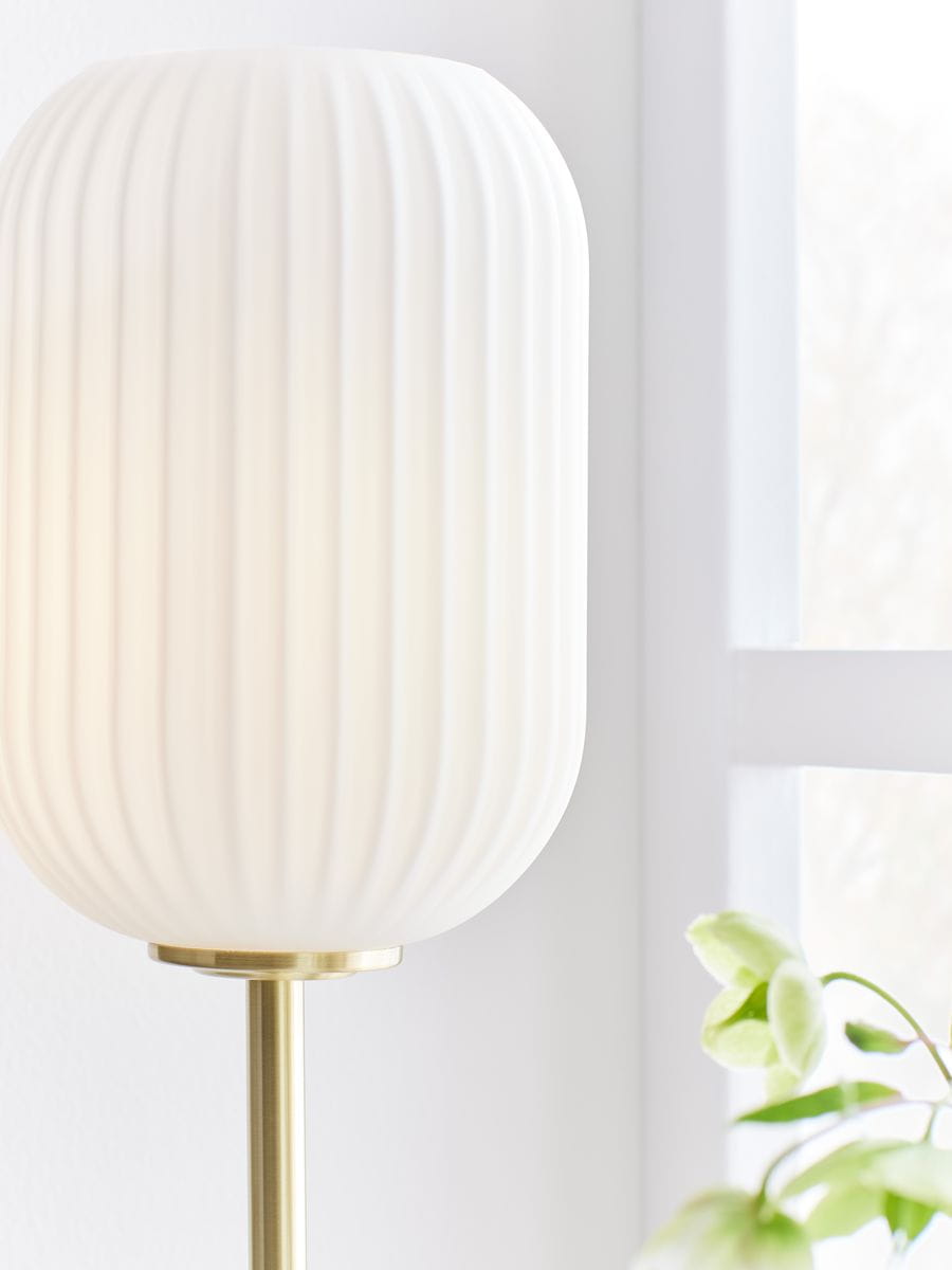 Lampa stołowa Cava z białym kloszem o żebrowanej fakturze, zamontowana na szczotkowanej mosiężnej podstawie. Ujęcie z boku, ukazujące detale materiału oraz kształt klosza. W tle widoczna roślina.