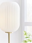 Lampa stołowa Cava z białym kloszem o żebrowanej fakturze, zamontowana na szczotkowanej mosiężnej podstawie. Ujęcie z boku, ukazujące detale materiału oraz kształt klosza. W tle widoczna roślina.