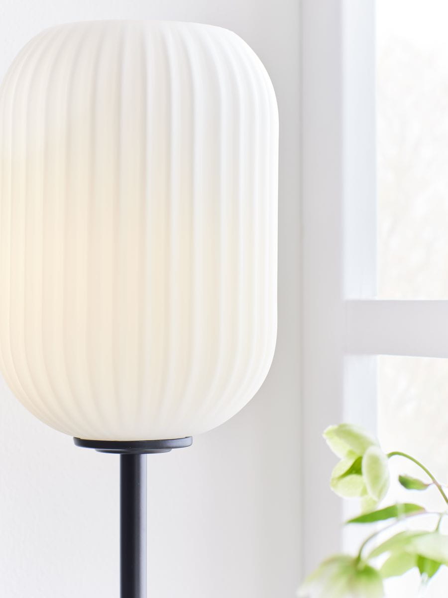 Lampa stołowa Cava 1-punktowa w kolorze czarnym i białym, z okrągłym, teksturowanym kloszem w odcieniu białym na smukłej, czarnej podstawie. Ujęcie z boku, z widocznymi detalami klosza i rośliną w tle.