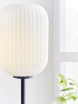 Lampa stołowa Cava 1-punktowa w kolorze czarnym i białym, z okrągłym, teksturowanym kloszem w odcieniu białym na smukłej, czarnej podstawie. Ujęcie z boku, z widocznymi detalami klosza i rośliną w tle.