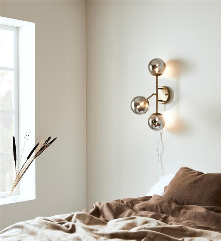 Trinity kinkiet 3-punktowy w kolorze mosiądzu z dymionymi kloszami. Ułożony na ścianie w aranżacji sypialni, z trzema kulistymi źródłami światła. Tło jasne, z oknem i dekoracyjnymi roślinami obok łóżka.