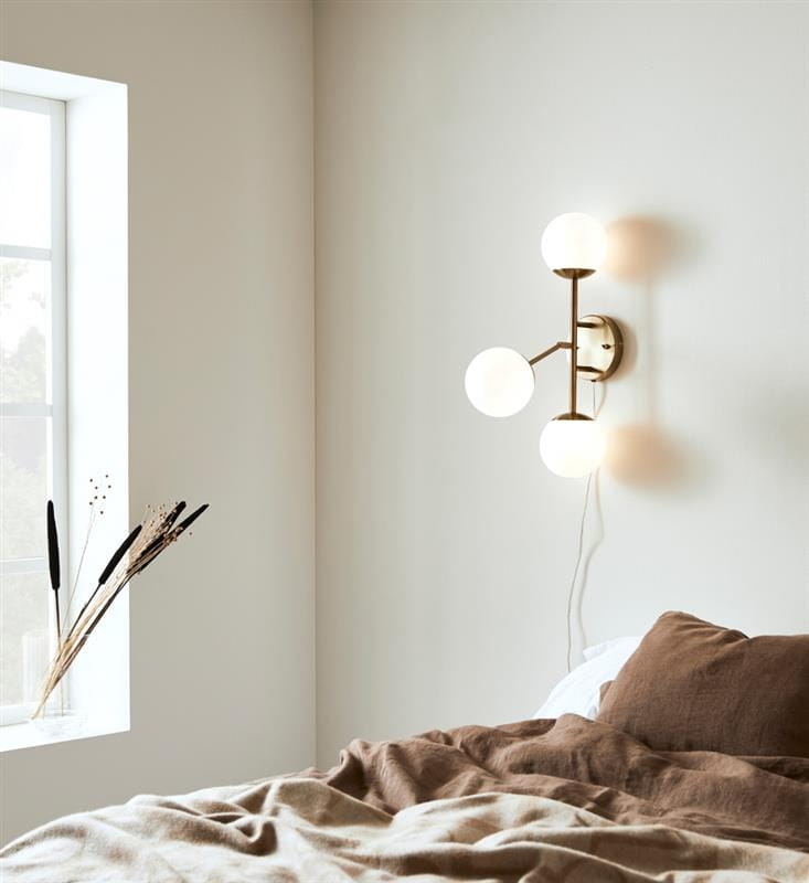 Kinkiet Trinity 3-punktowy w kolorze mosiądzu i białym, z trzema okrągłymi kloszami, zamontowany na jasnej ścianie obok okna. W tle widoczna pościel w odcieniach brązu, a na parapecie kilka dekoracyjnych traw.