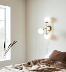 Kinkiet Trinity 3-punktowy w kolorze mosiądzu i białym, z trzema okrągłymi kloszami, zamontowany na jasnej ścianie obok okna. W tle widoczna pościel w odcieniach brązu, a na parapecie kilka dekoracyjnych traw.