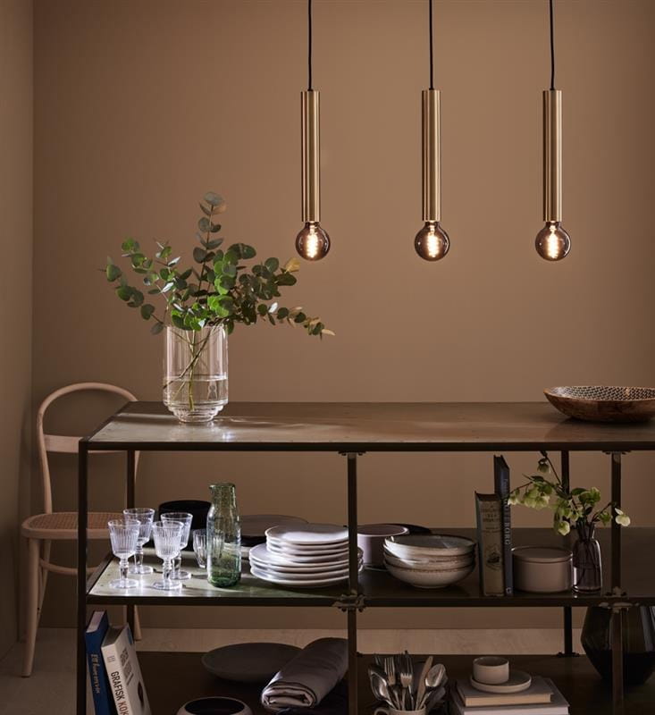 Lampa wisząca Sencillo 3-punktowa w kolorze mosiężnym, z trzema cylindrycznymi kloszami zawieszonymi na czarnych przewodach. Widoczny bukiet roślin w wazonie na stole, na którym znajdują się naczynia i szkło. Tło w ciepłym odcieniu.